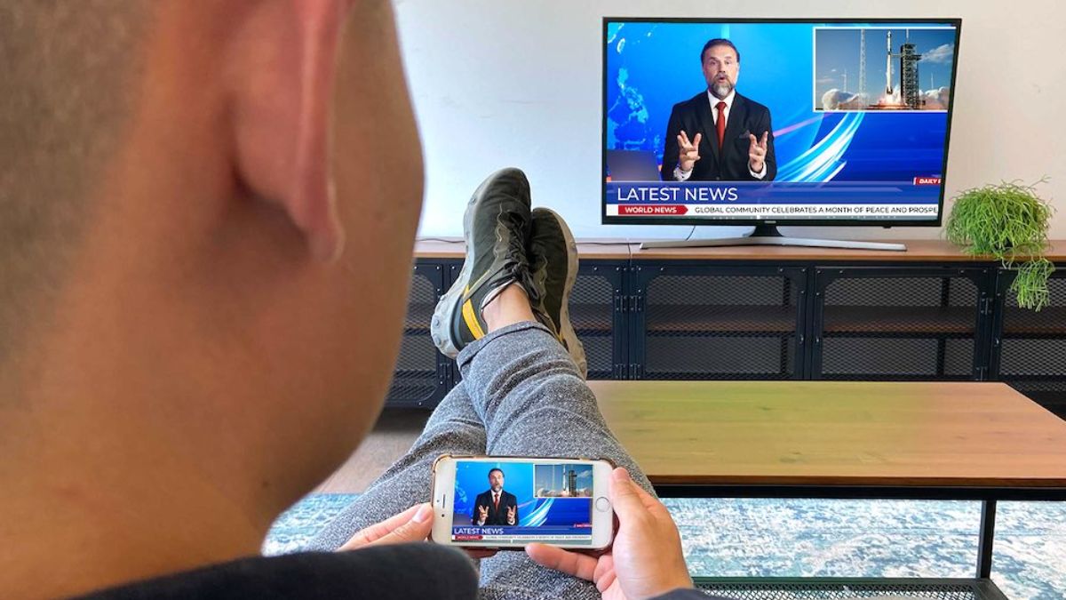 Người đàn ông nằm thư giãn, xem tin tức trên TV trong khi điện thoại cũng hiển thị nội dung tương tự, cho thấy tính năng screen mirroring giúp chia sẻ màn hình điện thoại lên TV lớn để xem thoải mái.
